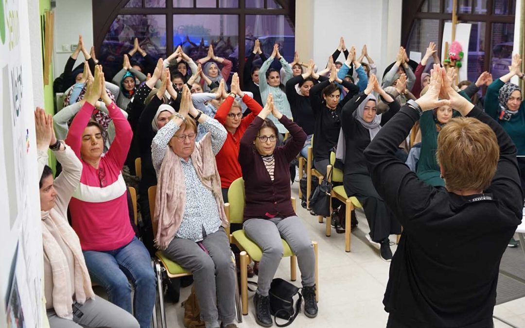 Etwa 20 Frauen und Mütter sind zu sehen. Sie sitzen im Publikum und halten die Arme in Yoga-Grußhaltung nach oben.