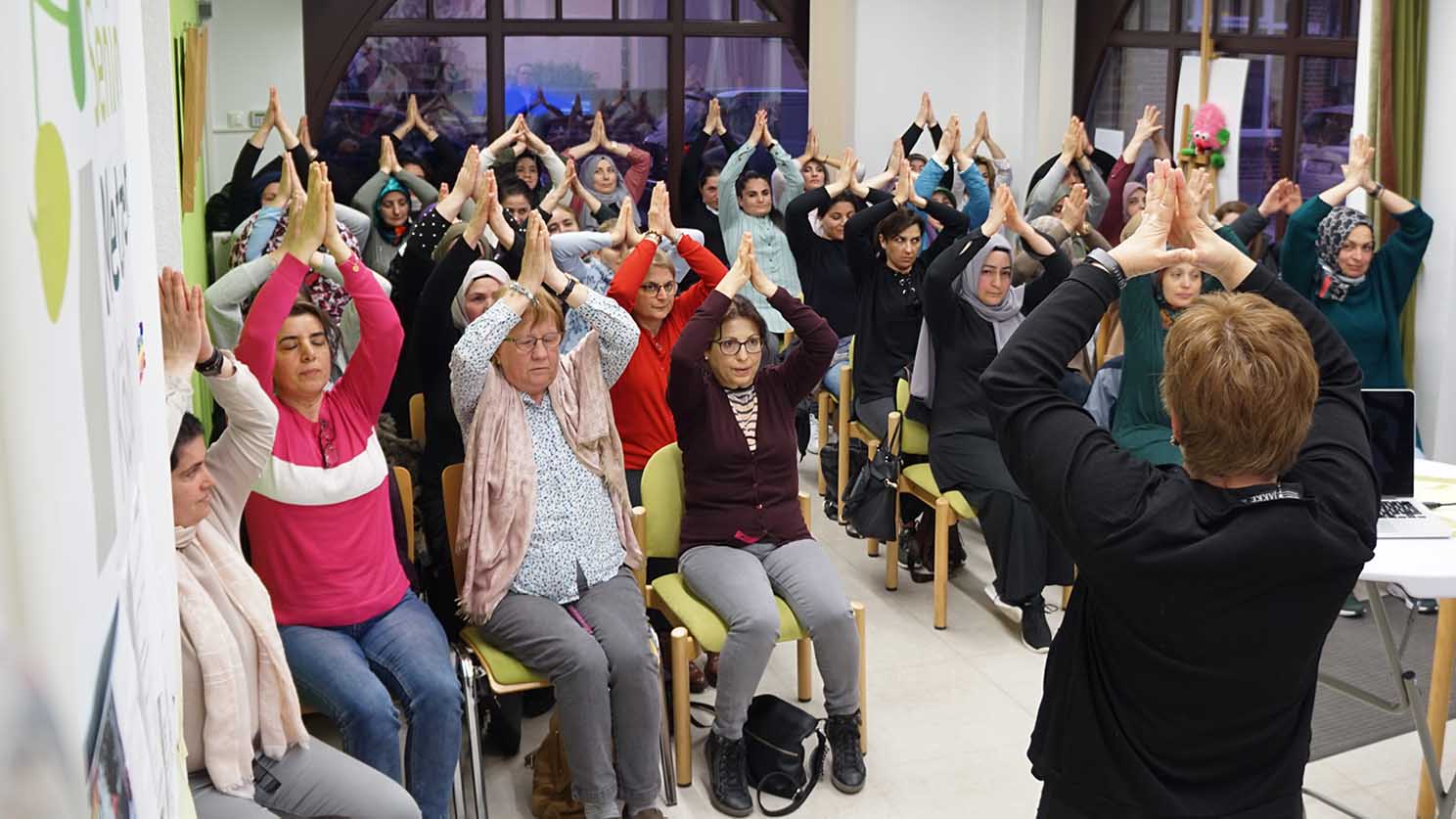 Etwa 20 Frauen und Mütter sind zu sehen. Sie sitzen im Publikum und halten die Arme in Yoga-Grußhaltung nach oben.